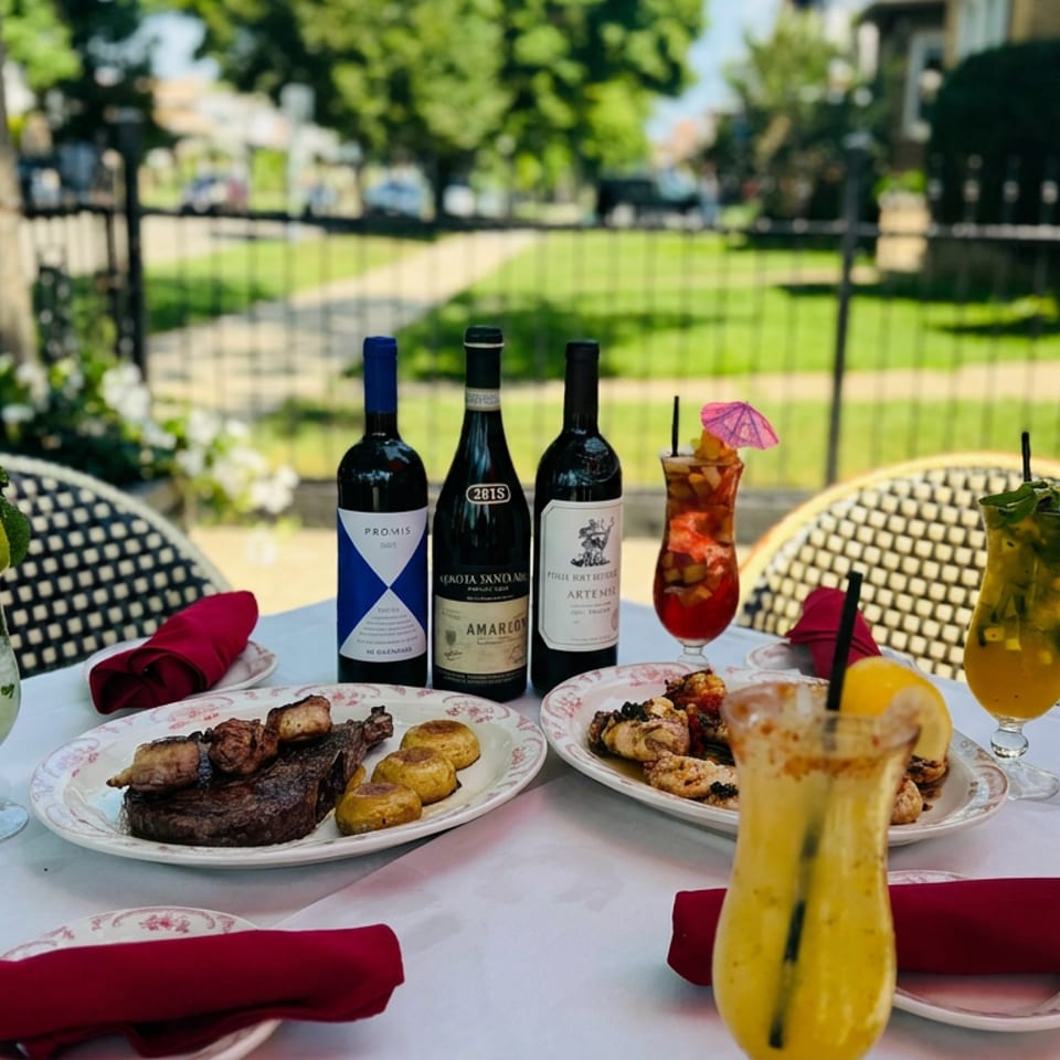 Outdoor restaurant table featuring plated meats and potatoes, wine bottles, colorful cocktails, and red napkins beside a sunny garden.
