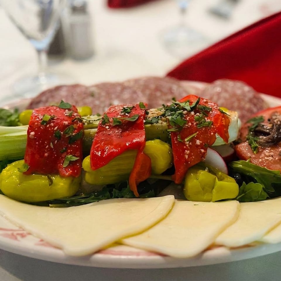 Plate of marinated red peppers, green vegetables, tomato, leafy greens, sliced cheese, and bread at a restaurant table.