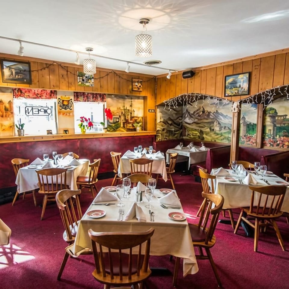 Warm, wood-paneled restaurant dining room with white tablecloths, set tables, burgundy carpeting, and colorful mountain murals.