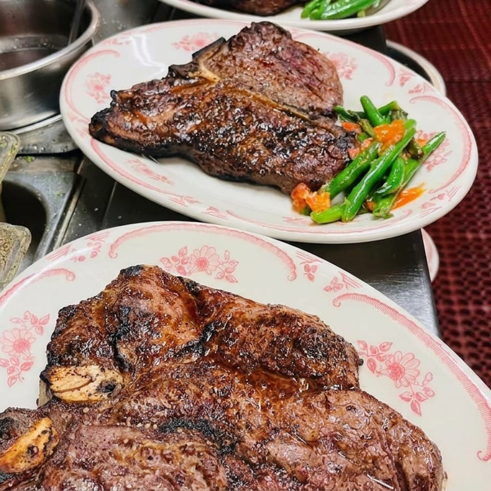 Grilled steaks on floral plates with green beans and carrots beside seasoning containers on a restaurant counter.
