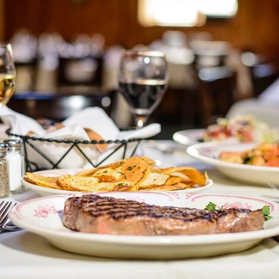 Restaurant table featuring grilled steak, sliced flatbread, plated dishes, wine glasses, and a bottle in a warmly lit dining room.